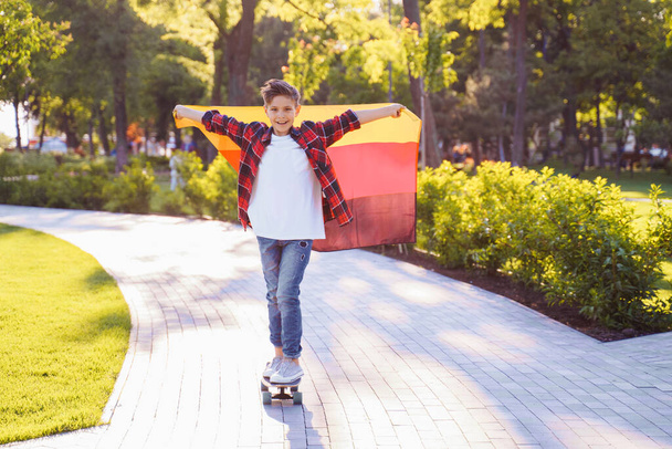 Guy of 9 years old riding skateboard on sunny road in the evening in the city park. Holding fluttering flag of Germany. Happy German day. German language, European education. - Photo, Image