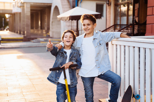 two attractive teenagers of Caucasian nationality 7 and 9 years old standing on sunny evening street in the city. Laughing, pointing fingers forward, holding scooter and skateboard. - Foto, imagen