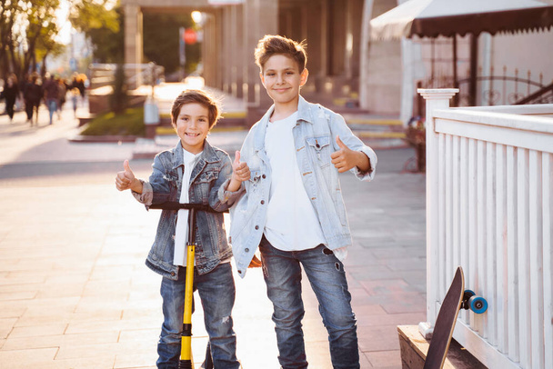 двое привлекательных подростков кавказской национальности 7 и 9 лет стоят на солнечной вечерней улице города. Смеясь, указывая пальцами вперед, держа скутер и скейтборд
. - Фото, изображение