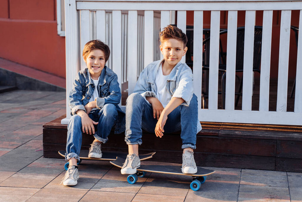 δύο ελκυστικοί έφηβοι Καυκάσιας εθνικότητας 7 και 9 ετών που στέκονται στην ηλιόλουστη οδό το βράδυ στην πόλη. Γέλιο, δείχνοντας τα δάχτυλα προς τα εμπρός, κρατώντας σκούτερ και skateboard. - Φωτογραφία, εικόνα