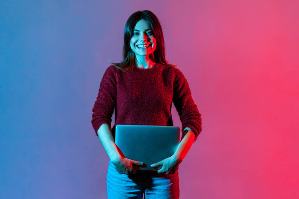 Portrait of successful happy office worker, pretty girl in casual pullover standing, holding closed laptop or folder, looking at camera with toothy smile, positive expression. neon light, studio shot - Fotografie, Obrázek