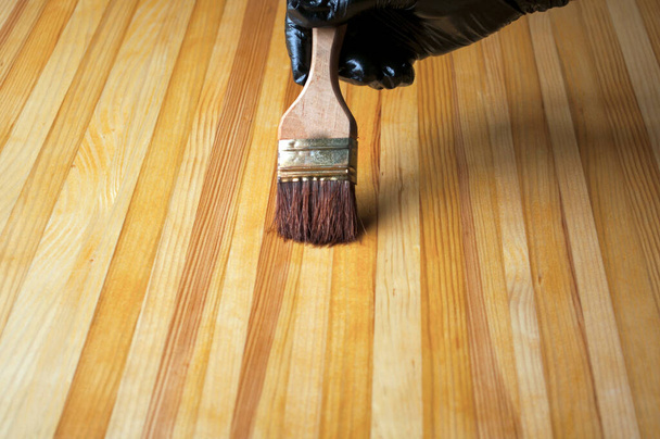 Hand in Hand malt ein schwarzer Handschuh. Malen mit dem Pinsel auf einem Holztisch. Behandlung einer Holzoberfläche zum Schutz gegen Alterung, Feuchtigkeit, Schmutz.  - Foto, Bild