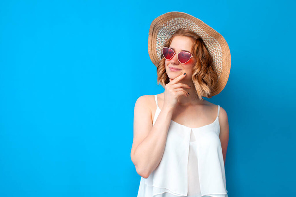 νεαρή στοχαστική κοπέλα σε ένα καπέλο ήλιο και γυαλιά σκέφτεται σε ένα μπλε απομονωμένο φόντο, γυναίκα σε καλοκαιρινά ρούχα, έννοια του καλοκαιριού - Φωτογραφία, εικόνα