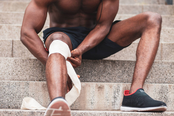 koncepce poranění kolena, afro Američan si obvazuje nohu obvazem, muž si při tréninku poranil koleno, zblízka - Fotografie, Obrázek