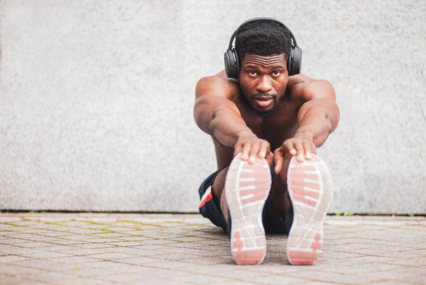 Αφροαμερικάνος αθλητικός τύπος μπαίνει για σπορ στην ύπαιθρο χωρίς μπλουζάκι στον τοίχο, ένας αθλητής με ακουστικά κάνει ζέσταμα, αντιγράφει χώρο - Φωτογραφία, εικόνα