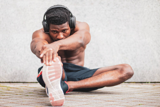 afrikanisch-amerikanischer Athlet treibt Sport im Freien ohne T-Shirt an der Wand, der Athlet mit Kopfhörer macht ein Workout, kopiert Platz - Foto, Bild