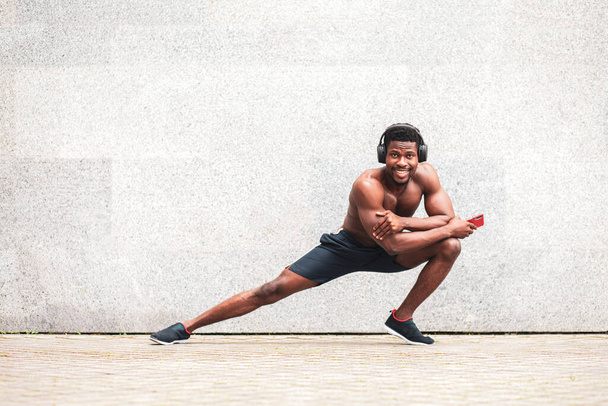 Afrikkalainen amerikkalainen urheilija treenaa ulkona seinää vasten, urheilullinen mies treenaa ilman t-paitaa ja kuuntelee musiikkia, kopioi tilaa - Valokuva, kuva