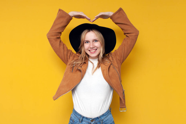 young girl in autumn clothes shows a heart with her hands and smiles on a yellow isolated background - Photo, Image
