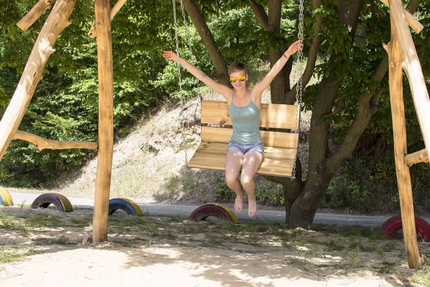 Une fille raccourcie en T-shirt et short, balançant sur une balançoire en bois, sans se tenir les mains et pieds nus - Photo, image