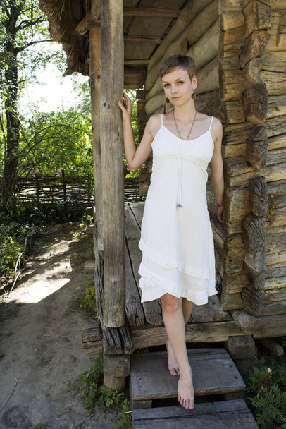 Egy fehérruhás, rövidvágású lány áll mezítláb egy öreg parasztház fából készült lépcsőjén. - Fotó, kép
