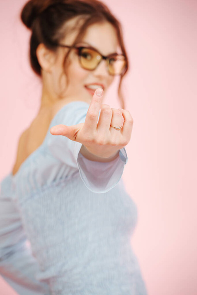 Cute eccentric provocative girl looking at camera, inviting with a finger. Over pink background. She wears glasses, two buns on her head and a shirt with an open top. Focus on a hand. - Foto, imagen