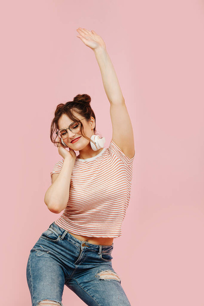 Emotional stilish woman in glasses dancing, while listening to headphones. She's wearing striped shirt and jeans. Her hair in two buns. - Фото, зображення