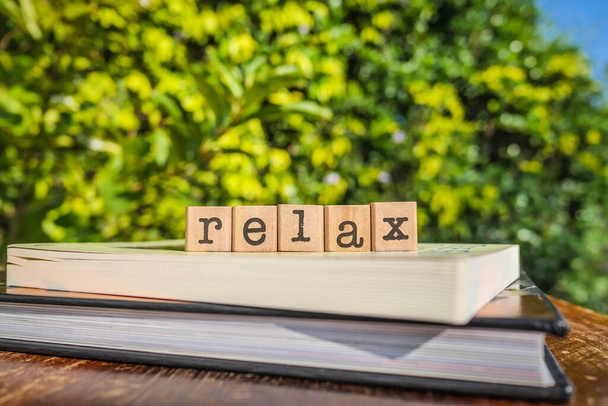 Слово RELAX, літери на кубиках блоків зверху книг та дерев'яний стіл з зеленими деревами, фон на природі. Поняття спокою, відпочинку та самодопомоги цитата
. - Фото, зображення