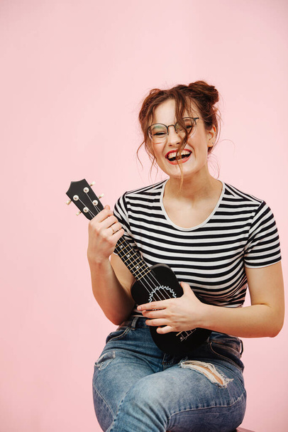 Lauging playful eccentric woman in glasses with black ukulele over pink background. She's wearing striped shirt and jeans. Her hair in two buns. - 写真・画像
