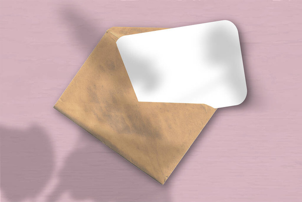 Una busta con un foglio di carta bianca strutturata sullo sfondo rosa. Mockup con una sovrapposizione di ombre vegetali. La luce naturale getta ombre dai gerani. Orientamento orizzontale. - Foto, immagini