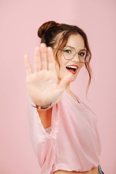 Eccentric overly happy young woman in glasses with two buns on her head over pink. She's making stop gesture, blocking camera. - Photo, image