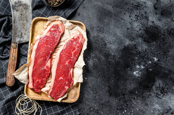 Rohe Roastbeef-Steaks auf einem hölzernen Tablett mit einem Fleischspalter. Marmorrind. Schwarzer Hintergrund. Ansicht von oben. Kopierraum. - Foto, Bild