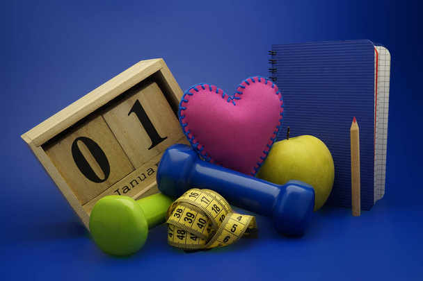 Starting healthy lifestyle on the first day of the year concept. Still life with dumbbells, measuring tape, calendar and new notebook on blue background - Photo, Image