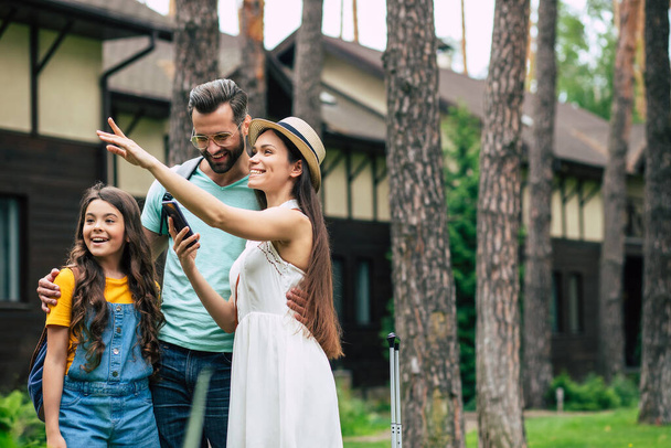 Attractions. A photo of a united happy family on a summer weekend staying outdoor near hotel, excited daughter looking the way mother pointing with her hand, smiling father looking at phone. - Photo, image