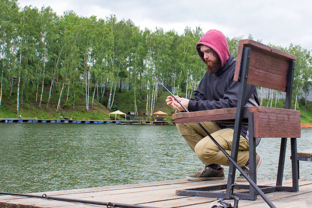 суворий норвезький чоловік сидить на причалі й ловить рибу рибалкою. бородатий носить чорну куртку і червоний каптур на голові.. - Фото, зображення