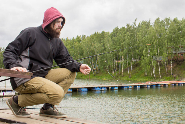 ノルウェー人の男性がドックに座って釣竿で釣りをしています。髭を生やした男は黒いジャケットと赤いフードを頭につけている. - 写真・画像
