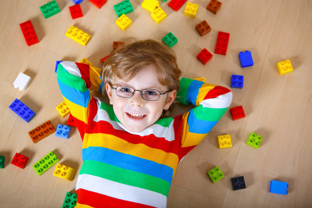 Malé blonďaté dítě s brýlemi si hraje se spoustou barevných plastových bloků uvnitř. Kluk nosí barevné tričko a baví se s budováním a tvorbou - Fotografie, Obrázek