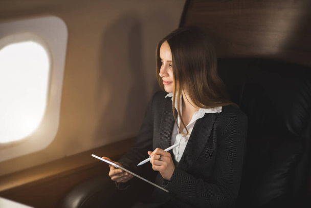 Nice young businesswoman in formal attire, with makeup, looking at screen of tablet with satisfied expression, working remotely while flying on private jet. Business, traveling and technology. - Photo, Image