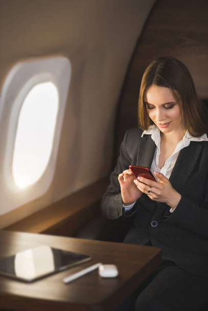 Beautiful young businesswoman traveling in private jet. Brown haired lady in white blouse, formal suit holding smartphone, sitting in comfortable seat in airplane. - 写真・画像