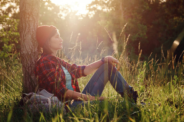 Young hipster girl enjoy sunset at forest. Travel woman with backpack. Enjoying nature sunset. Relaxing in park at sunrise. Trekking concept - Zdjęcie, obraz
