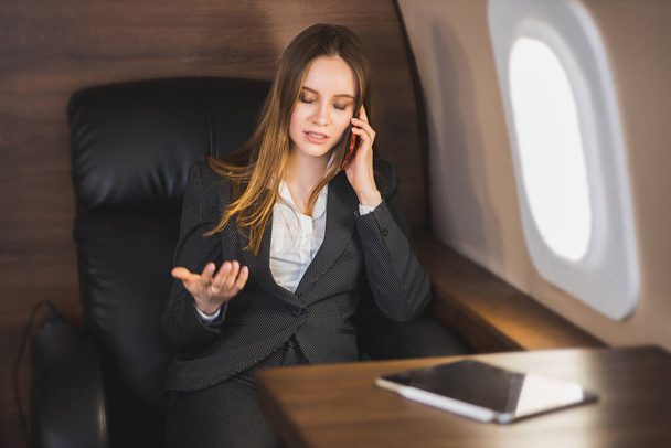 Attractive nervous young caucasian businesswoman is talking on the phone with an irritated facial expression, gesturing, sitting on a private plane. Business, emotions concept. - Fotografie, Obrázek