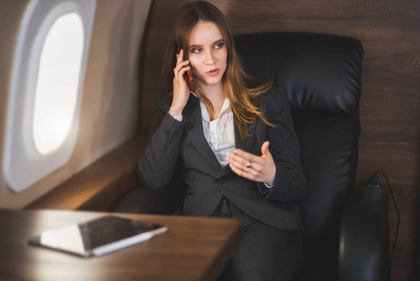 Cheerful happy brown haired business lady in white blouse and formal suit, sitting in comfortable seat, laughing talking on mobile phone during flight on first class plane. - Foto, imagen