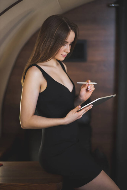 Lovely slender young woman wearing sexy black dress in private jet. Successful young woman entrepreneur holding digital tablet, working during the flight. Technology concept. - Fotó, kép