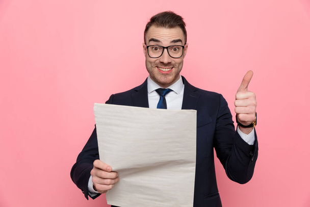happy elegant guy in suit wearing glasses and reading newspaper, making thumbs up sign and smiling, standing on pink background - Фото, зображення