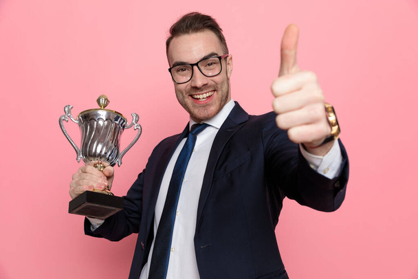 heureux jeune homme d'affaires en costume portant des lunettes, tenant trophée et faisant signe pouces vers le haut, debout sur fond rose - Photo, image