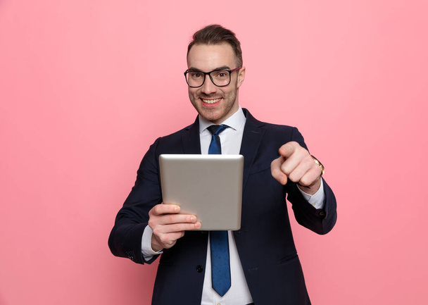 šťastný mladý podnikatel v obleku nosí brýle, drží kartu a čtení e-mailů, ukazuje prstem a stojí na růžovém pozadí - Fotografie, Obrázek