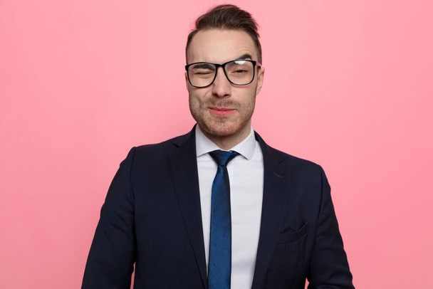 sexy young businessman in suit wearing glasses and winking, standing on pink background - Foto, imagen