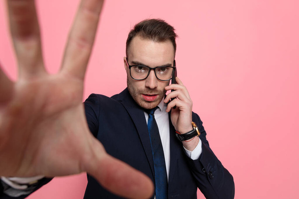 молодий елегантний чоловік у костюмі в окулярах, розмовляє по телефону і обрамляє, стоїть на рожевому фоні
 - Фото, зображення