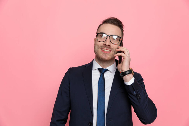 happy young man in suit wearing glasses, talking on the phone and looking up, standing on pink background - Photo, image