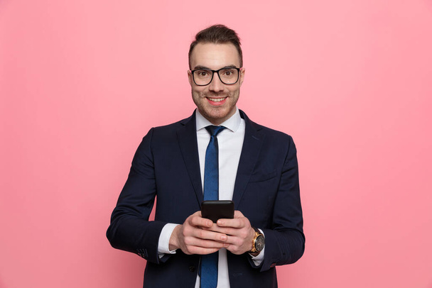 χαρούμενος νεαρός επιχειρηματίας με κοστούμι φορώντας γυαλιά, χαμογελώντας και γράφοντας μηνύματα, στέκεται σε ροζ φόντο - Φωτογραφία, εικόνα