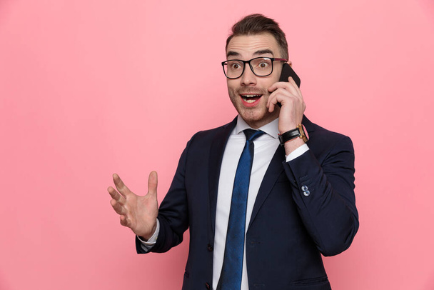 счастливый элегантный мужчина в костюме разговаривает по телефону, удивленный новостями, жестом и глядя в сторону, стоя на розовом фоне
 - Фото, изображение
