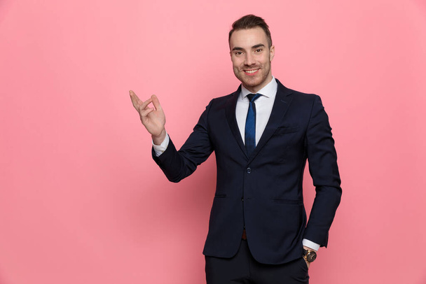 happy young elegant man in suit presenting to side and smiling, holding hand in pocket and standing on pink background - Foto, immagini