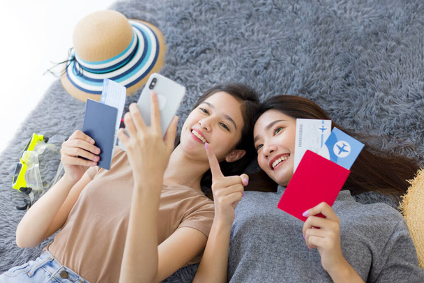 Dwie młode Azjatki leżące na podłodze, trzymające paszporty i bilety podczas korzystania z telefonu w poszukiwaniu atrakcji turystycznych. Ładne dziewczyny planują wakacyjną wycieczkę w lecie w domu. - Zdjęcie, obraz