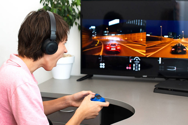 十代の男の子がオンラインでヘッドフォンとジョイスティックでコンピュータゲームをプレイし、ゲームコンソール. - 写真・画像