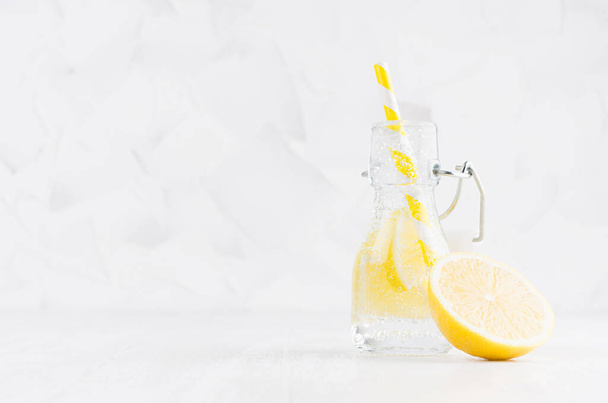 Refreshing cold fruit drink with citrus lemon, bright bubbles and straw in transparent yoke bottle in elegant white interior on wood table, copy space. - 写真・画像