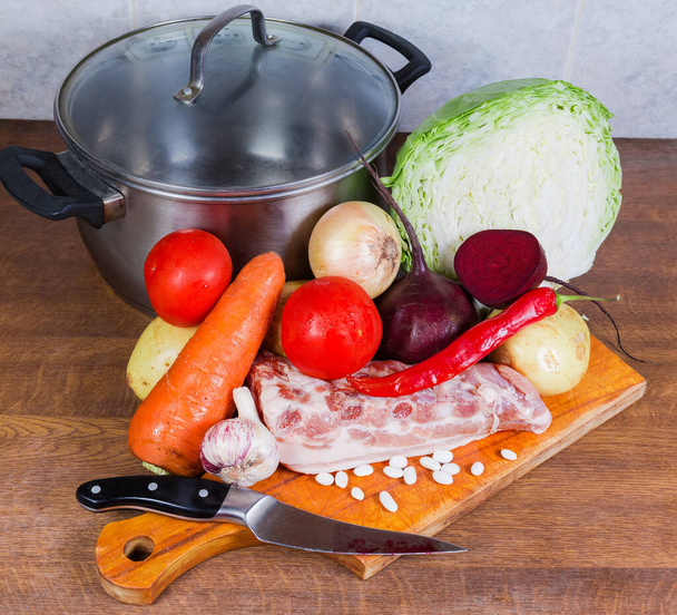 Обычное сырье для приготовления типичного борща из красной свеклы - свиные ребрышки, свежие овощи и бобы, пустой горшок, нож на кухонном столе
 - Фото, изображение