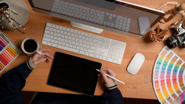 Overhead shot of female designer working with digital tablet, mock-up computer and designer supplies on wooden desk  - 写真・画像