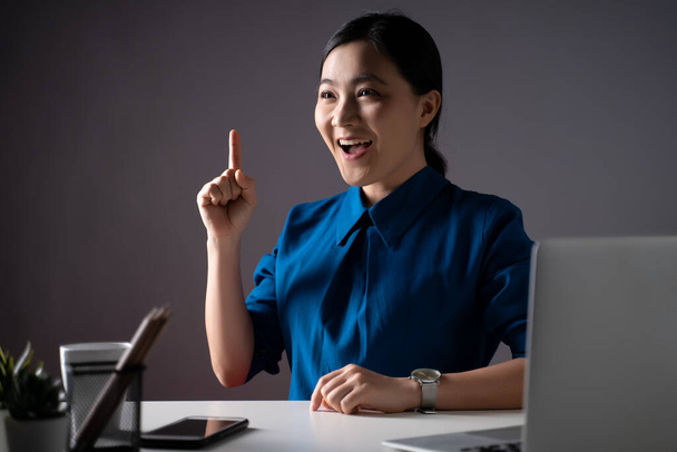 Azjatka szczęśliwa uśmiecha się w niebieskiej koszuli wskazując palcem, pracując nad laptopem w biurze. odizolowany od przeszłości. Niski klucz. - Zdjęcie, obraz
