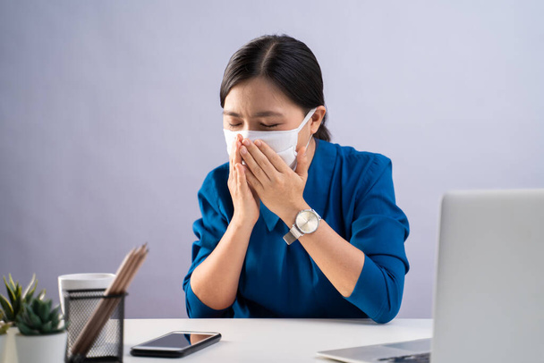 Mujer asiática en camisa azul con mascarilla protectora, estaba enferma de dolor de garganta, trabajando en un portátil sentado en la oficina. aislado sobre fondo blanco
. - Foto, imagen