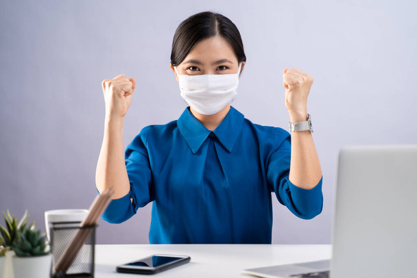 Femme asiatique en chemise bleue portant un masque protecteur montrant son poing faire un geste gagnant et de travailler sur un ordinateur portable au bureau. isolé sur fond blanc. - Photo, image