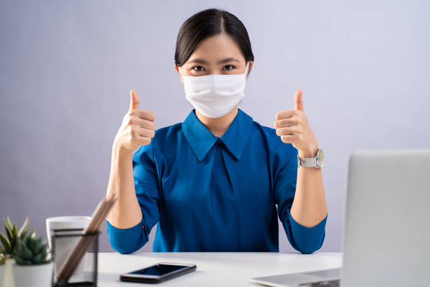 Asian woman in blue shirt wearing protective face mask happy and showing thumb up and working on a laptop at office. isolated on white background. - Foto, afbeelding